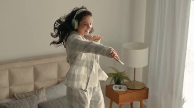 Kafkasyalı genç enerjik kadın esmer yatakta dans ediyor yatak odasında rahat kulaklıklar takıyor eğlenceli kulaklıklar takıyor müzik dinliyor hafta sonu akıllı telefon hobileri söylüyor heyecanlı zıplama eğlencesi aktif
