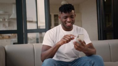 Mutlu, kaygısız, Afrikalı, Amerikalı erkek, rahat bir ev sahibi, koltukta cep telefonu kullanıyor, sosyal medya alışverişi yapıyor, gülüyor, cep telefonu akıllı telefonu interneti kullanıyor.