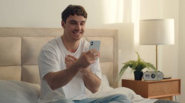 Kafkasyalı mutlu bir adam yatak odasında oturmuş elinde cep telefonu ile görüntülü konuşma blogu çekiyor internet konferansı, sosyal ağlar, sosyal ağlar, konuşan iş yeri hoparlörleri.
