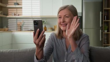 Mutlu kaygısız, yaşlı, Kafkasyalı, yaşlı, yaşlı, yaşlı, emekli, videolu, el sallayarak selam veren bir gülümseme. Uzaktan iletişim kuran yaşlı kadın. Cep telefonu uygulamalı kanepe.