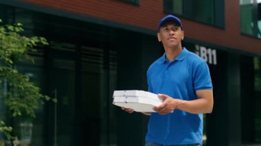 Açık havada yürüyen Afro-Amerikan kurye elinde pizza kutusu pizzacısı erkek teslimatçı teslimat servisi hazır yemek lokantası karton paket şehir postacısının iş yürüyüşü müşteriye gidiyor.