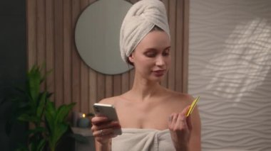 Banyo havlusuna sarılı beyaz bir kadın çevrimiçi bayan ev satın alıyor kredi kartı alıyor cep telefonu internet e-ticaret alışverişini kullanarak online veri ödeme mağazasına giriyor.