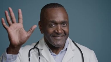 Gülümseyen Afrikalı Amerikalı doktor kameraya bakıyor. Stüdyonun mavi arka planında sağlık hizmetlerini selamlıyor. Sağlık desteğini selamlıyor.