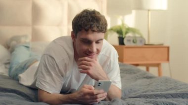 Kafkasyalı bir adam yatağında uzanmış cep telefonu kullanıyor gülümseyen adama mesaj atıyor erkek iletişimi online internet teknolojisi sosyal medya ağları yatak odaları sohbet aletleri.
