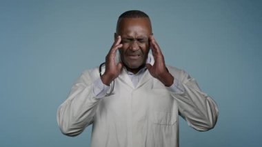 Mutsuz Afro-Amerikan erkek doktor erkek sağlık çalışanı mavi stüdyoda tıbbi görevli baş ağrısı çekiyor kafa hastalığı masaj tapınaklarında kronik migren ağrısı çekiyor
