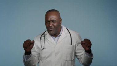 Komik, mutlu, Afro-Amerikan erkek doktor, erkek, sağlık çalışanı, doktor, stüdyo mavi arka planda zafer dansı yapan doktor, sağlık sigortası reklamını kutluyor.