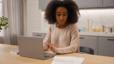 Afro-Amerikalı kadın serbest çalışan iş kadını mutfak masasında evraklarla çalışıyor. Ciddi kız internet faturalarını ödüyor. Ev bilgisayarında yerel bütçe internet hizmetleri.