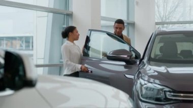 Afrikalı Amerikalı satıcı kadın tüccar müşterinin yeni modern elektrikli otomobil adamı seçmesine yardımcı oluyor. Araba alan kadın menajer ile konuşuyor. Müşteri satışı aracı kiralama servisi alıcısı oto kuaföründe oturuyor.
