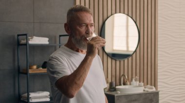 Yaşlı Kafkasyalı yaşlı, yaşlı, olgun, emekli erkek, banyo banyosunda sabah su içiyor, camları tutuyor, sağlıklı alışkanlıkları dengeliyor. H2O ferahlatıcı, susuz, yaşlı nesil sağlık hizmetleri.