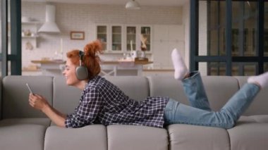 Mutlu bayan rahat koltukta uzanıyor cep telefonuyla sohbet ediyor sosyal medya akıllı telefon uygulaması kablosuz kulaklıkla beyaz kadın kız evdeki kulaklıklarda müzik dinliyor.