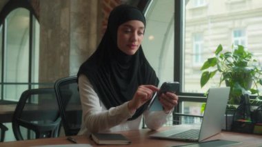 Müslüman kadın tesettüre girmiş Arap iş kadını. Masa başında çalışan gülümseyen iş kadını. Cep telefonu bağımlısı.