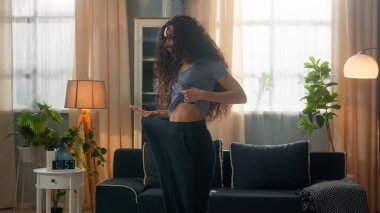 Mutlu, şaşkın, gülümseyen, etnik İspanyol kadın, yaşlı kot pantolon giyen, zayıf kadın, kilo kaybı, spor, beslenme, sağlıklı bir oturma odası.