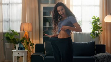 Gülümseyen Latin kökenli, mutlu, sporcu, kadın sporcu, büyük kot pantolon giyen, ince bel kaslı, kilo kaybı yaşayan bir kadın.