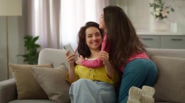 Mutlu aile hafta sonları birlikte. Beyaz kadın anne cep telefonu kullanıyor aktif küçük kız çocuk okul kızı kız çocuğu oturma odasındaki kanepeye atlayıp anneye sarılıyor.