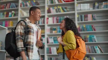 İki Afrikalı Amerikalı erkek Latin kadın kütüphane kütüphanesinden konuşuyor edebiyat öğrencileri kitapçıdan kitaplar okuyor iletişim kuruyor kadın üniversite çalışmalarını sırıtıyor.