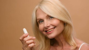 Mature middle-aged elderly woman model makeup hygienic lipstick applying moisturizing lip balsam natural beauty face skin care anti-aging cosmetic flirty senior lady smiling on beige studio background
