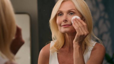 50 'li yaşlarda, güzel, olgun bir kadın yüzüne pamuk yastıkla toz ve lüks kozmetik uyguluyor. Kusursuz pürüzsüz cilt makyajı yapıyor. Aynalı ev banyosunda, yüz kırışıklıkları, yaşlanmayı önleyen güzellikler.