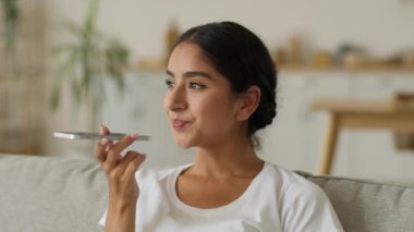 Evde gülümseyen mutlu Hintli kadın Arap pozitif Arap bayan bayan oturma odasında cep telefonuyla konuşan akıllı telefon mikrofon konuşan hoparlör kaydı yapan sesli mesajlar kaydeden.