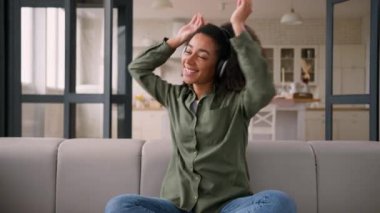 Neşeli, kaygısız kız Afrikalı Amerikalı genç bayan ev sahibi kanepeye oturup dans eden eller müzik kulaklıkları, müzik, müzik, müzik, kablosuz kulaklıklar, dans, rahatla.