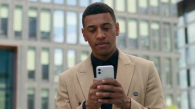 Düşünceli Afro-Amerikan etnik iş adamı şehir dışında ofis binası işveren işadamı erkek girişimci akıllı telefon sohbetleri yapan müşteriyi düşünerek kayan cep telefonuna göz atıyor.