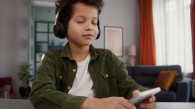 Afrikalı Amerikalı küçük çocuk kulaklık takıyor cep telefonu uygulaması kullanıyor müzik dinliyor evde dans eden müzik dinliyor eğlence dansı eğitimi molası