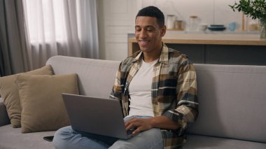 Mutlu gülümseyen Afrikalı Amerikalı etnik adam evde çalışıyor kanepe görevlisi dizüstü bilgisayarı kullanarak bilgisayar kullanıyor internet oyunu oynuyor uygulama alışverişi yapıyor.