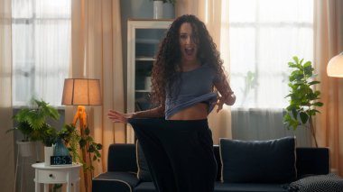 Mutlu, şaşkın, gülümseyen, etnik İspanyol kadın, yaşlı kot pantolon giyen, zayıf kadın, kilo kaybı, spor, beslenme, sağlıklı bir oturma odası.