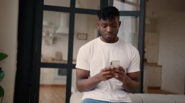Afro-Amerikalı erkek ev sahibi kiracı erkek arkadaş cep telefonu uygulamasını evde tek başına kullanıyor. Bekar erkek kullanıcı üzgün üzgün. İnternetten cep telefonuyla mesajlaşırken hayal kırıklığına uğramış.