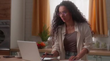 İspanyol kadın Latin kadın mutfakta dizüstü bilgisayarda çalışıyor. Alışveriş notları yazıyor. Baş ağrısı çekiyor.