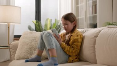 Gadget bağımlılığı. Neşeli, pozitif, beyaz çocuk, reşit olmayan kız çocuk gülümsüyor cep telefonuyla yazılar yazıyor sosyal medyayı besliyor küçük çocuk sanal oyunlar oynuyor, internette kanepede sohbet ediyor.