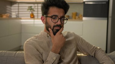 Düşünceli Hintli, düşünceli, etnik bir adam ev mutfağında kafası karışmış zor bir karar verme sorununu çözüyor mutsuz bir erkek kanepede tek başına yatıyor derin düşüncelere dalmış şüpheli bir soru düşünüyor.