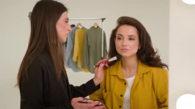 Beyaz kadın makyaj sanatçısı, stüdyo yansıması içinde son moda blog yazarı kadın stilistini pudralıyor. Etkilenen yüz fırçasına gölge görüntüsü uyguluyor. Kozmetik moda güzelliğini filme çekmeye hazırlanıyor.