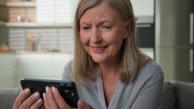 Mutlu son sınıf 60 'lar olgun beyaz kadın yaşlı büyükanne cep telefonu akıllı kadın emeklisi iyi haberler okuyor evde sosyal medyada olumlu mesajlar alıyor gülümsüyor.