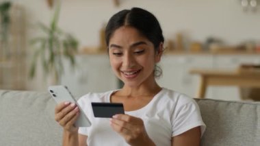 Mutlu gülümseyen Hintli kadın müşteri Arap kız elinde cep telefonu kredi kartı ödemesi çevrimiçi ödeme güvenli numara yetkilendirme satın alma ev alışveriş internet bankacılık