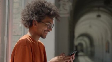 Üniversitede cep telefonu kullanan Afrikalı Amerikalı etnik erkek öğrenci akıllı telefondan gülümseyen mutlu sosyal medya bağımlısı eğitim programlarını internetten kırıyor.