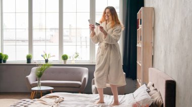 İnternet üzerinden rezervasyon yaptıran beyaz bir kız bornoz alışverişi yapan, cep telefonu takan, kredi kartı ödemesi yapan mutlu bir kadın yatak odasında dans ederek indirimi kutluyor.