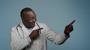 Memnun Afro-Amerikalı doktor başarılı bir tıbbi servis önerisi kliniği tavsiye etti. Stüdyonun mavi arka planında, sağlık hizmetleri reklamlarında yazı yazmak için boş bir alanı işaret etti.