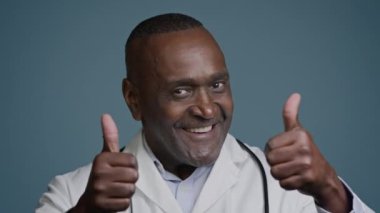 Beyaz önlüklü, gülümseyen Afrikalı Amerikalı doktor. Stüdyonun mavi arka planında baş parmak gösteriyor.