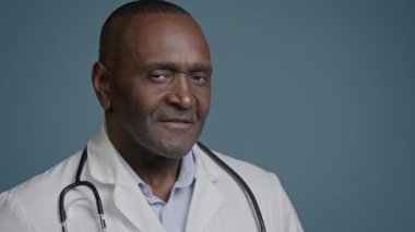 Yakın plan başarılı gülümseyen Afro-Amerikan erkek doktor sağlık çalışanı, portre uzmanı, kardiyolog, pediatrist dişçi, kamera görüntüsü mavi arka plan stüdyosunda.