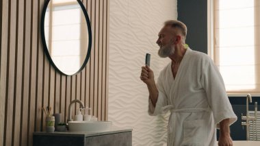 Evde bornozlu, neşeli, mutlu, yaşlı, komik, Kafkasyalı, yaşlı, yaşlı, saç tarağı taradıktan sonra saçlarını tarayan olgun bir adam.