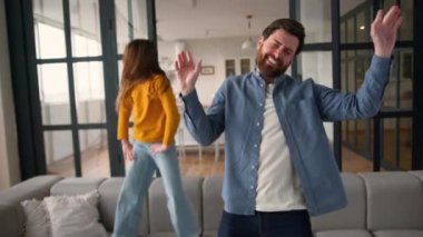 Olumlu Kafkas çocuk küçük kız kanepeye atlıyor erkek ebeveyn kaygısız aile müzik eşliğinde dans ediyor baba kız oynaşıyor. Çocuğun babası güler, şarkı söyler evde birlikte eğlenir, mutlu mesut eğlenir.