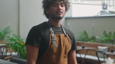 Kendinden emin gülümseyen mutlu Latin Amerikalı erkek mutlu kafeterya çalışanı önlüklü garson temizlikçi barista küçük işletme sahibi sokak kafeteryasındaki müşterilere bakarak el ele tutuşuyor.