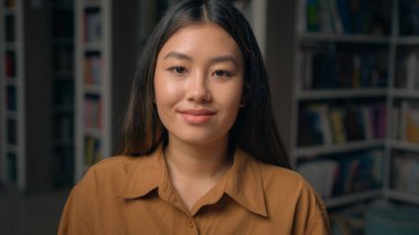 Close-up happy asian young woman posing in library looking at camera girl student sitting at desk with books read preparing for university exam studying homework book reading hobby moving footage blur