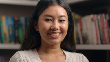 Kitaplığın yanındaki üniversite kütüphanesinde poz veren Asyalı kadın mutlu mutlu gülümseyen Çinli etnik kız öğrenci model müşterimiz içeride çekilmiş kadın portresine bakıyor.