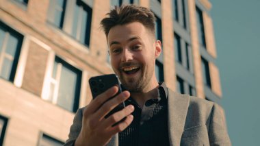 Mutlu Kafkas erkek işadamı online kazanan e-posta okuyucu iyi haberleri kontrol ediyor şehir dışında ofis merkezinin yakınında akıllı telefon kullanarak zaferi telefonla kutluyor.