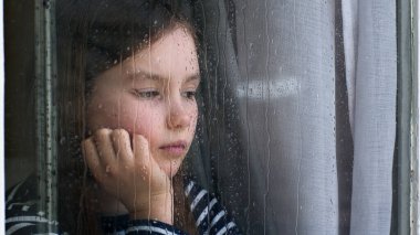 Kafkasyalı küçük kız pencereden dışarı bakıyor. Yağmurlu hava. Üzüntü, stres bunalımı, boşanma, çocuk, psikolojik travma.