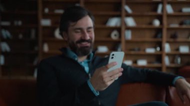 Şaşırtıcı derecede mutlu Kafkasyalı yetişkin erkek gülümseyen erkek işadamı cep telefonunu internet üzerinden kullanıyor sosyal ağda akıllı telefon kullanıyor geniş gülümsemeli sohbet yapıyor kapalı bir apartman dairesinde oyun kazanıyor