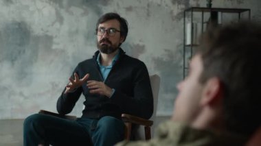 Olgun Kafkasyalı psikolog, gözlüklü erkek psikoterapist. Travma sonrası stres bozukluğunu iç mekandaki savaş kamuflajlı asker memuruna anlatıyor.