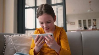 Neşeli, mutlu, beyaz bir çocuk cep telefonuna dokunuyor, akıllı telefon ekranı oynuyor, gülümsüyor, okulda zaferi kutluyor.