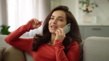 Mutlu, genç, beyaz bir kadın gülümseyen kadın, gülen kız, kullanıcı, iş kadını, neşeli ev sahibi, konuşan cep telefonu, akıllı telefon, sohbet eden, dedikodu yapan, iyi haberleri paylaşan, oturma odasında kanepe koltuğu tartışan.